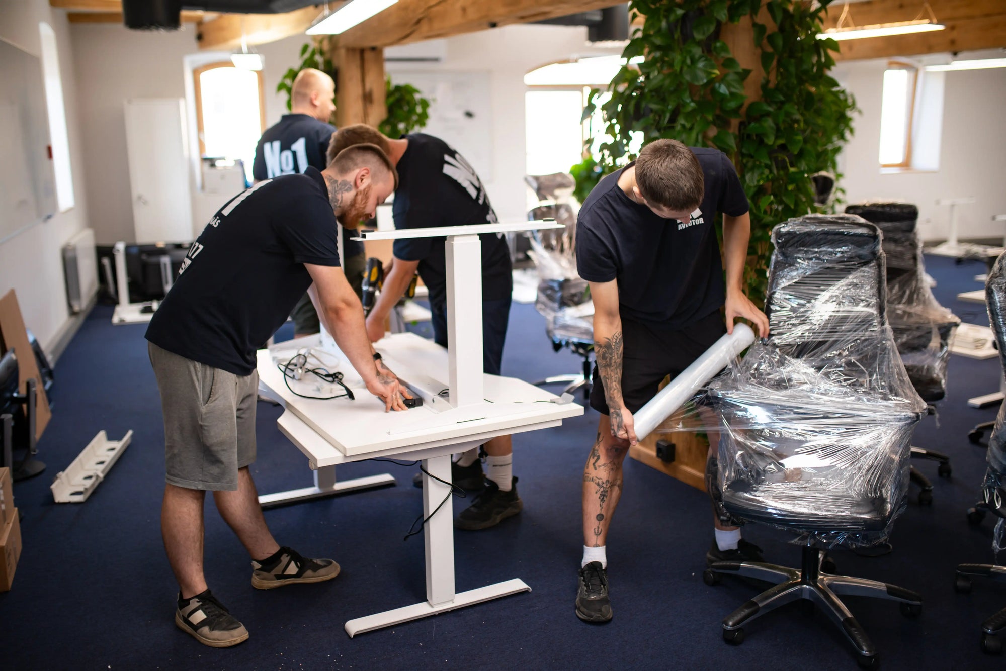 Three men working on assembling office chairs in a room with blue carpet and plants.